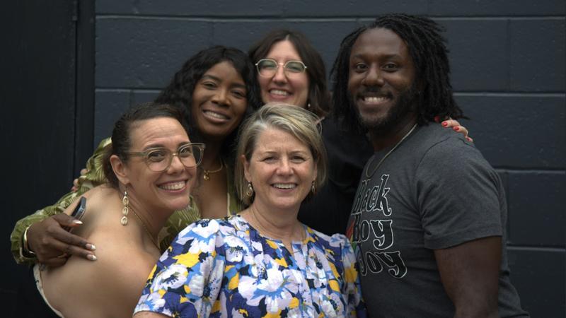 One Black woman, three white women and one Black man huddle together smiling for a group shot in front of a wall.
