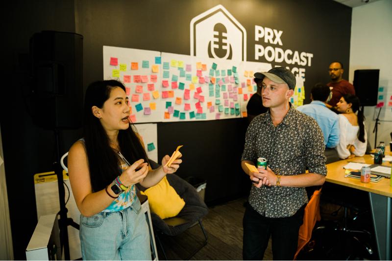 Two people speak in front of a wall of Post-It notes with the PRX Podcast Garage logo in the background.