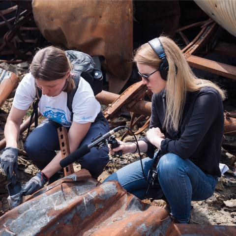A white woman with a microphone interviews a white woman in a war-torn location.
