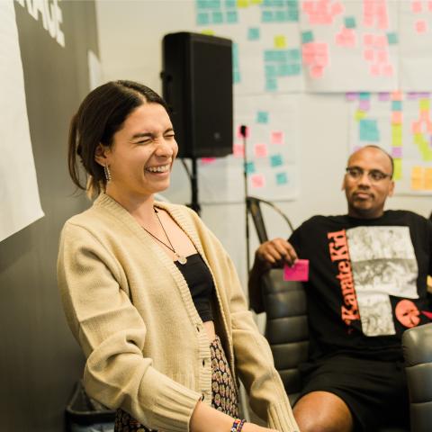 In the foreground a midshot of a woman with dark hair laughing and in the background two people sit with a wall of Post-its behind them.