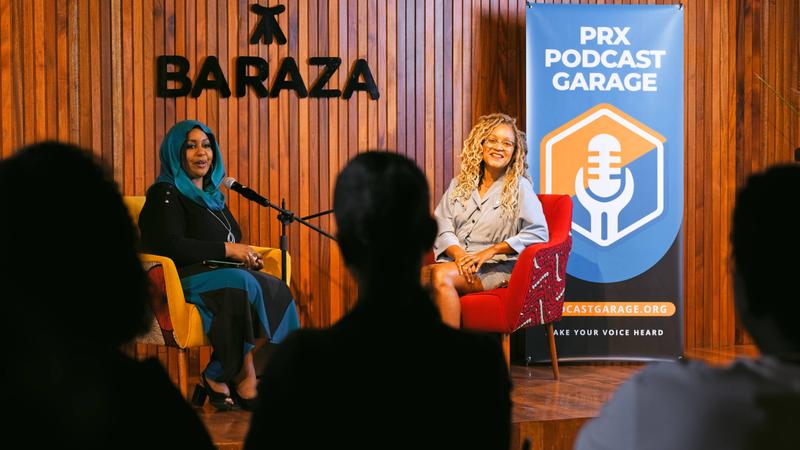 Two Black women, one wearing a headscarf, sit in chairs in front of a crowd with the PRX Podcast Garage and Baraza Media Lab logos behind them.