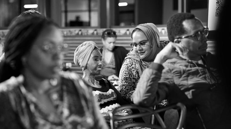 A black and white photo of two women wearing headscarves in the midground speak to each other.