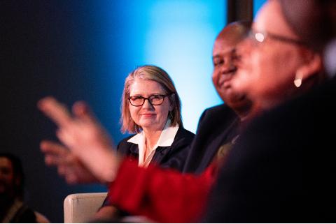 A white woman (Kerri Hoffman) with shoulder-length hair and glasses is seen in focus with a Black man (Glynn Washington) and white white woman (Catherine Burns)in the foreground.