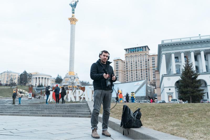 A white man dressed in black holding recording equipment stands in front of a monument in Kyiv, Ukraine.