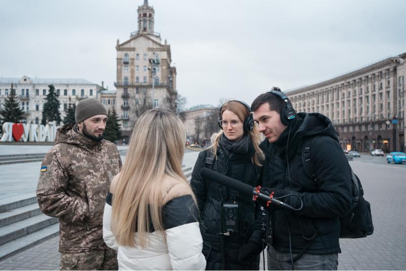 A white man and a white woman in black interview a white woman with blonde hair and a white man in military fatigues with a microphone on the streets of Kyiv, Ukraine.